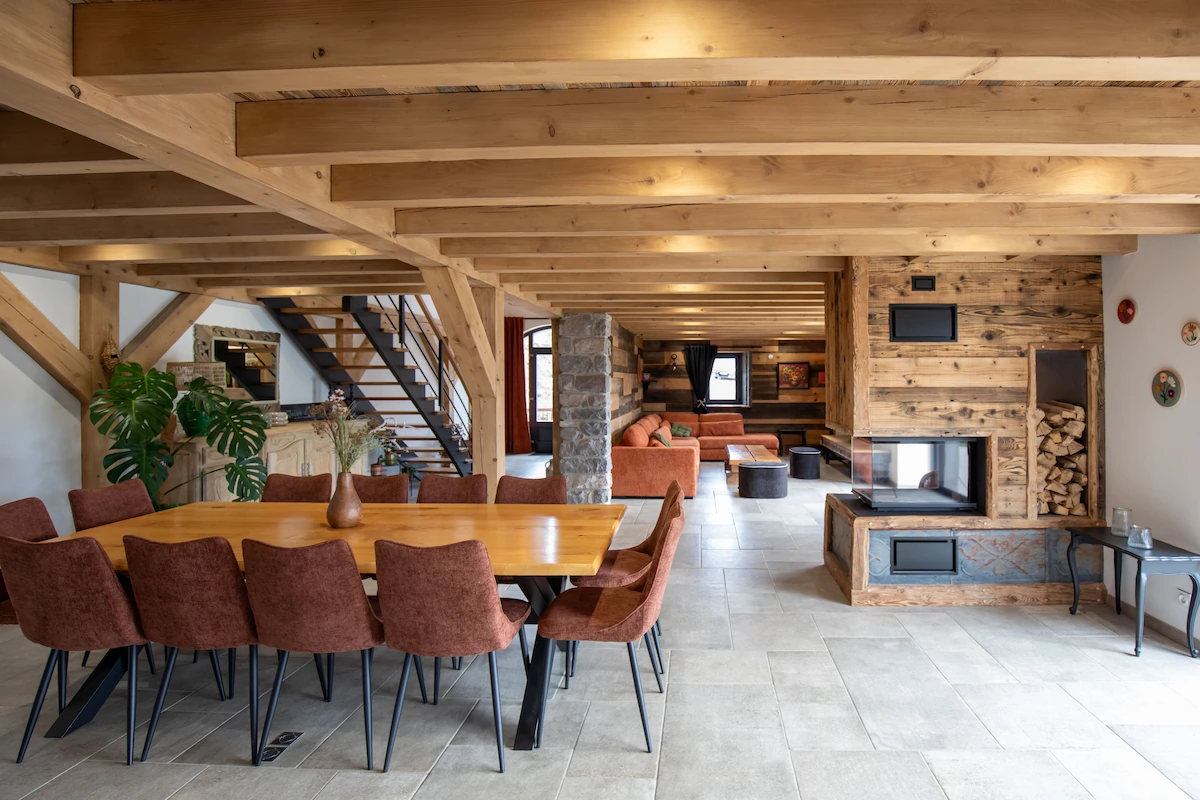 Rustic open living space with wooden beams, a dining table with brown chairs, a stone fireplace, and an orange sofa set.