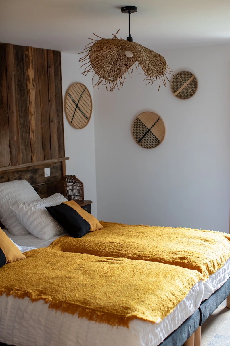 Bedroom with two single beds pushed together, white bedding, mustard yellow blankets, and black and mustard throw pillows under a woven pendant light and wall decorations.