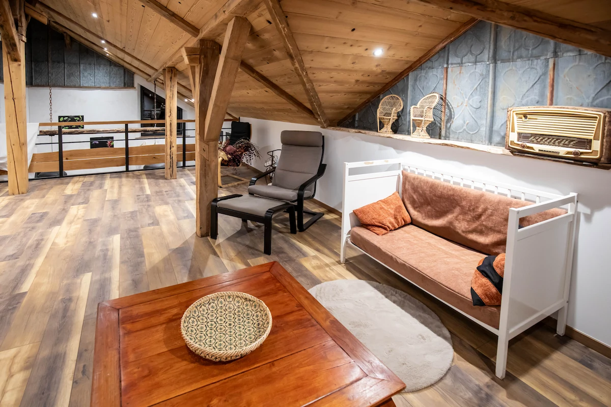 Cozy attic living space with wooden ceiling and floor, a gray armchair, white sofa with brown cushions, vintage radio, and a decorative basket on a wooden coffee table.