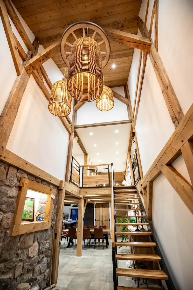 Interior of a rustic two-story room with wooden beams, stone wall, hanging wicker lamps, and a staircase leading to an open loft.