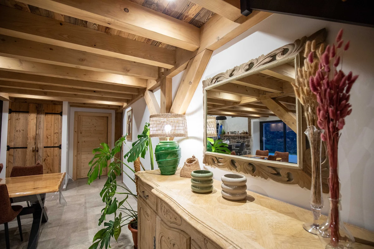 Rustic interior hallway with wooden beams, a sideboard with decorative items, a large mirror, and a dining table with chairs.