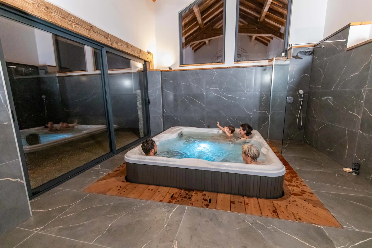 Four people relaxing in a modern indoor hot tub with gray marble walls and wooden ceiling.