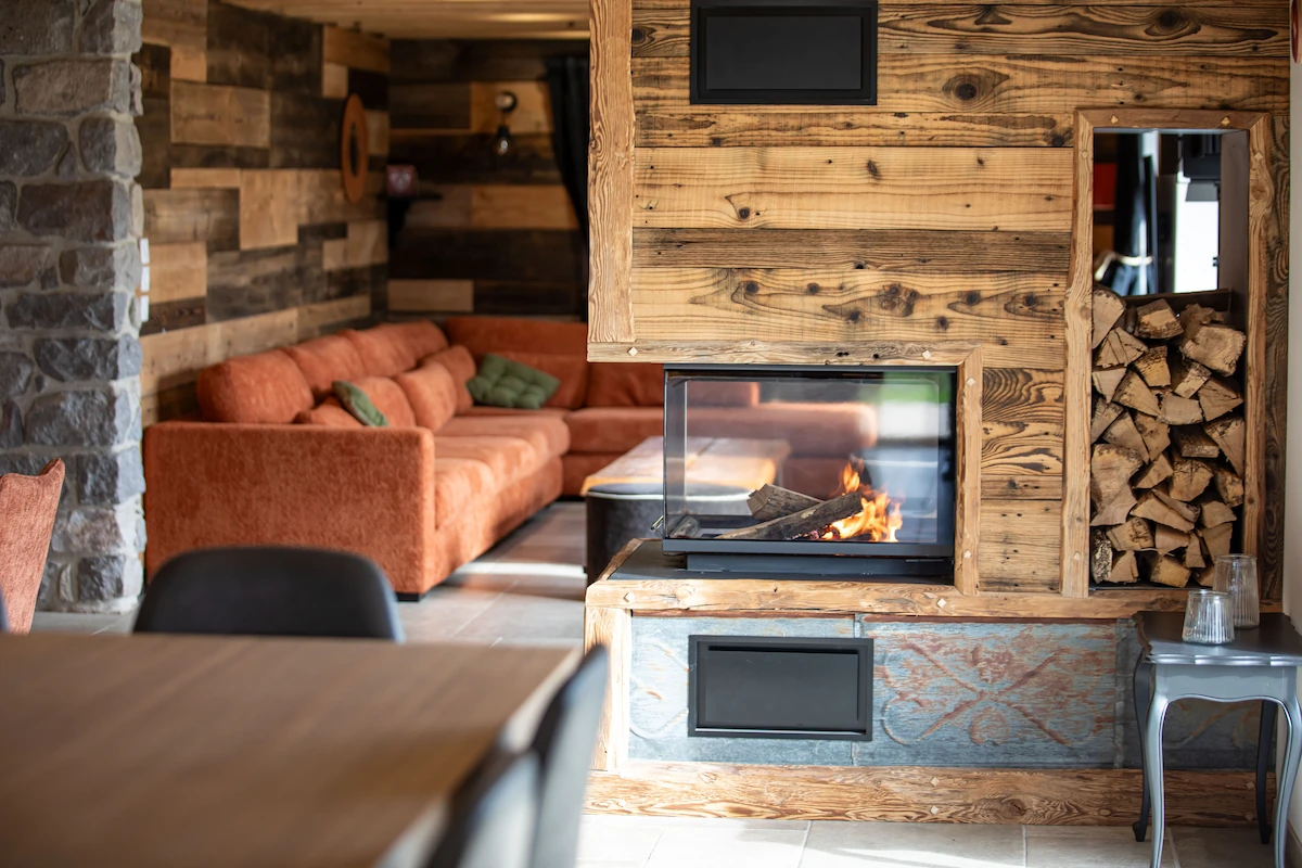 Cozy living room with a modern wood-paneled fireplace burning firewood, orange sofa, and stacked firewood storage.