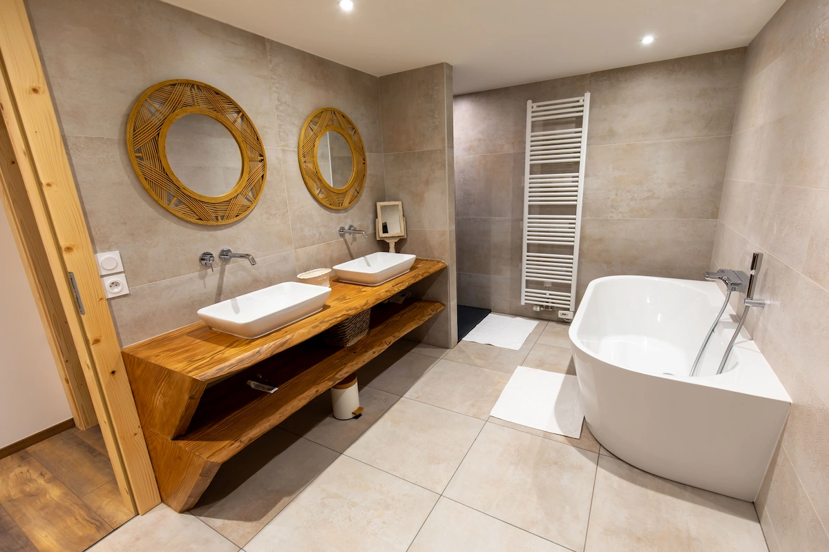 Modern bathroom with two rectangular white sinks on a wooden vanity, round rattan mirrors, a freestanding white bathtub, and beige tiled walls and floor.