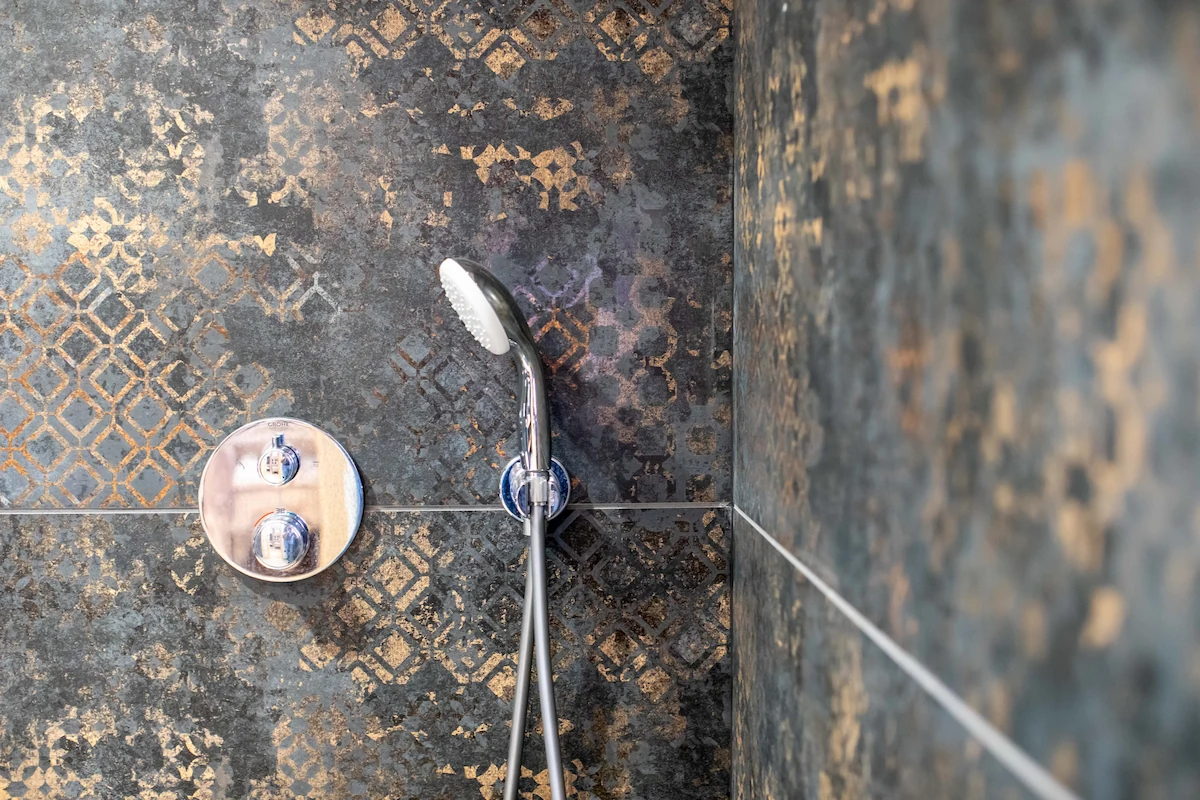 Chrome handheld shower head and control knobs mounted on dark patterned tiles in a shower.