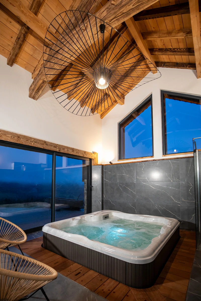 Indoor hot tub with bubbling water under a wooden vaulted ceiling with large windows and modern black wire-frame pendant light.