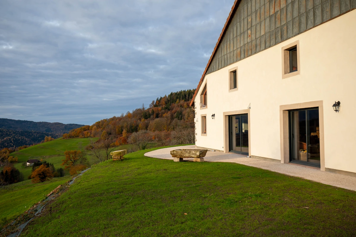 Modern house with large windows on a green hillside overlooking autumn-colored trees and distant hills under a cloudy sky.