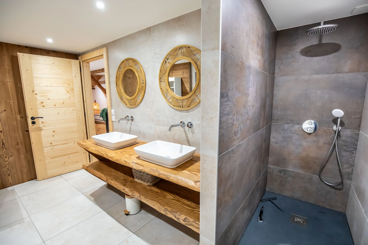 Modern bathroom with two white vessel sinks on a wooden countertop, round wicker-framed mirrors, and a walk-in shower with gray tiles and a rain showerhead.