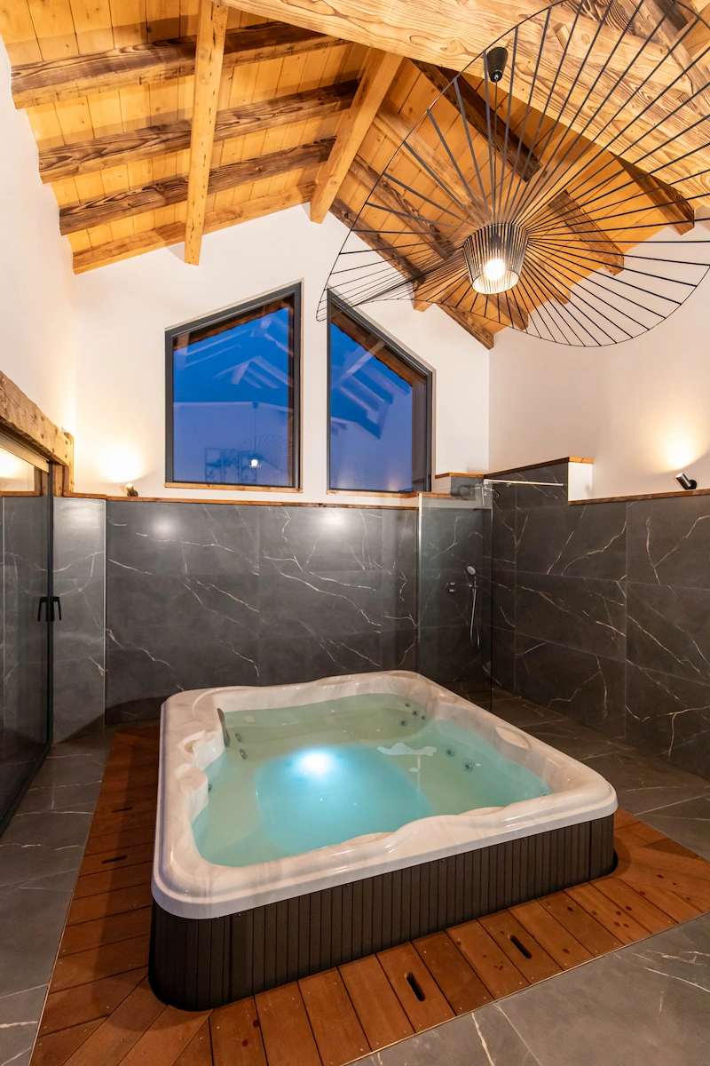 Indoor hot tub with illuminated water, surrounded by gray marble walls and wooden ceiling beams.