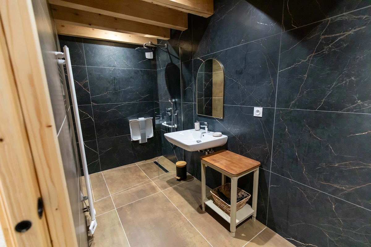 Modern bathroom with black marble walls, walk-in shower, wall-mounted sink, and wooden stool with basket underneath.