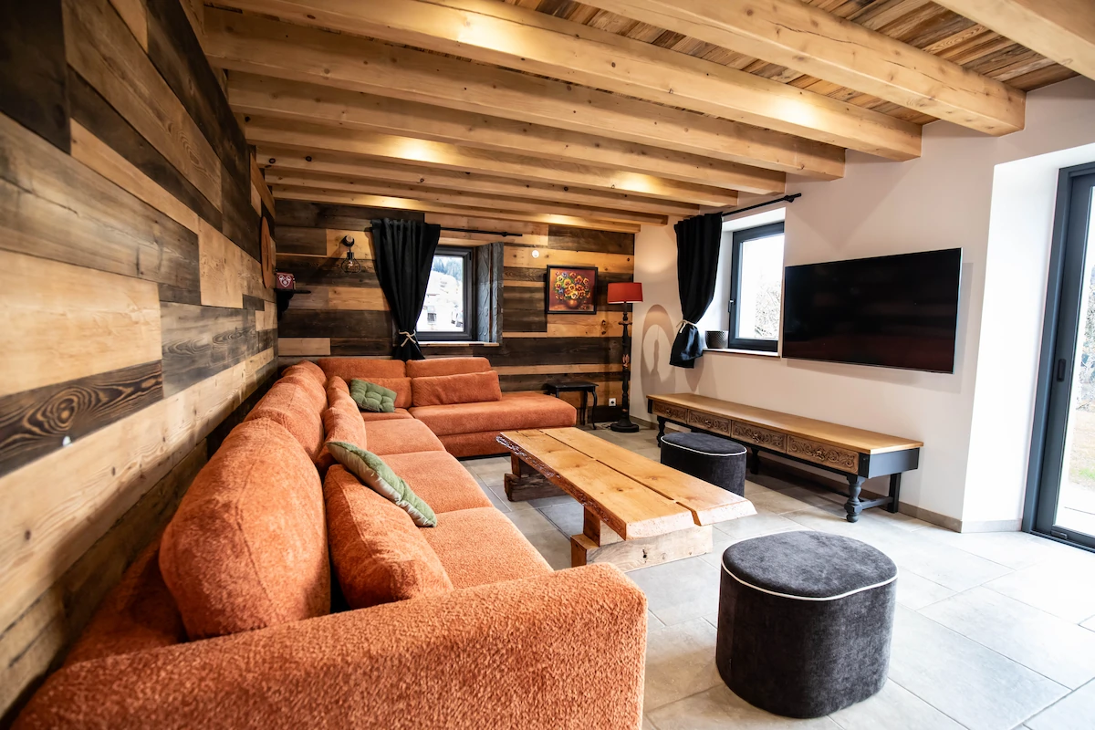 Cozy living room with wooden beam ceiling, orange sectional sofa, wooden coffee table, mounted TV, and black curtains on windows.