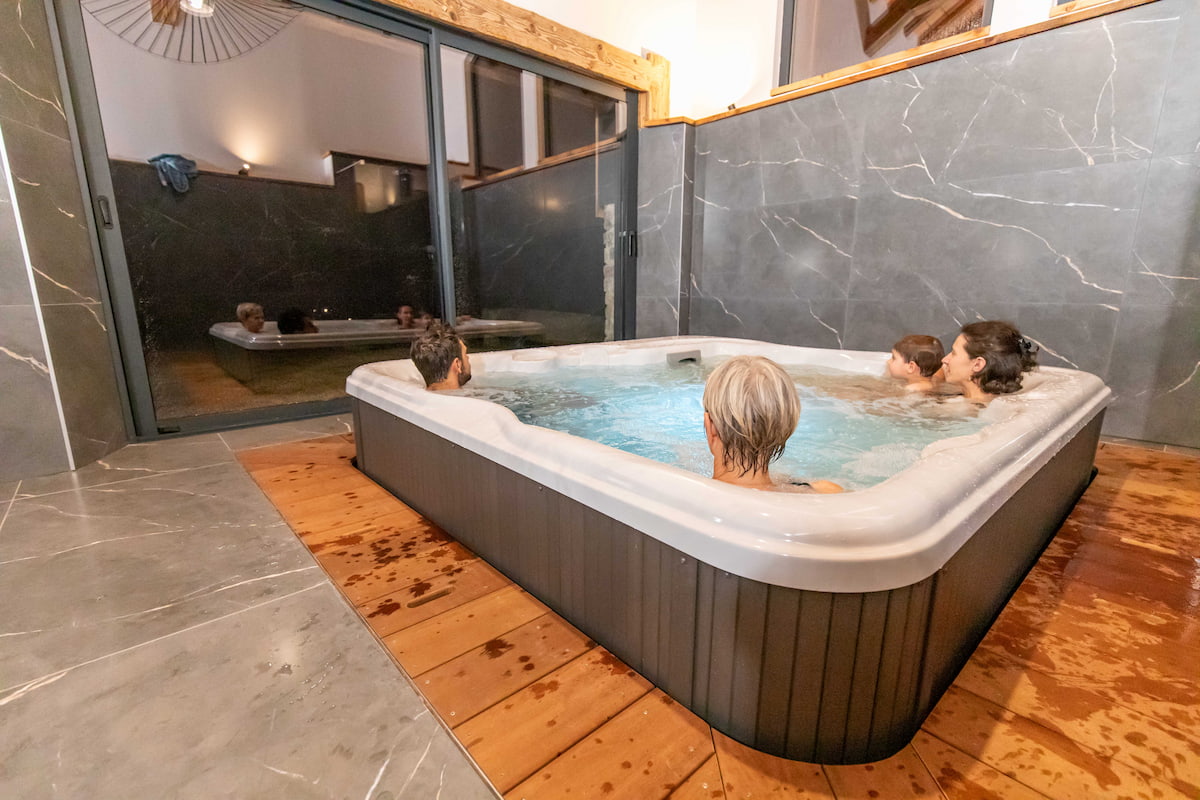 Four people relaxing in an indoor hot tub with gray marble walls and wooden flooring.