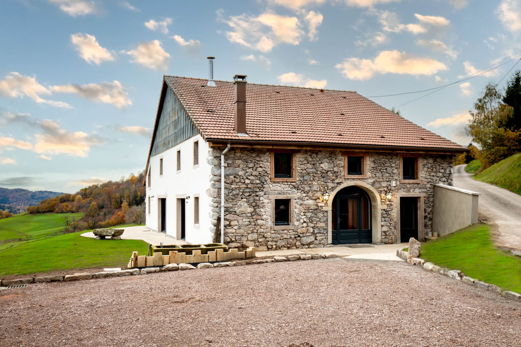 Stone and white house with a tiled roof surrounded by green grass and a gravel driveway under a blue sky with scattered clouds.