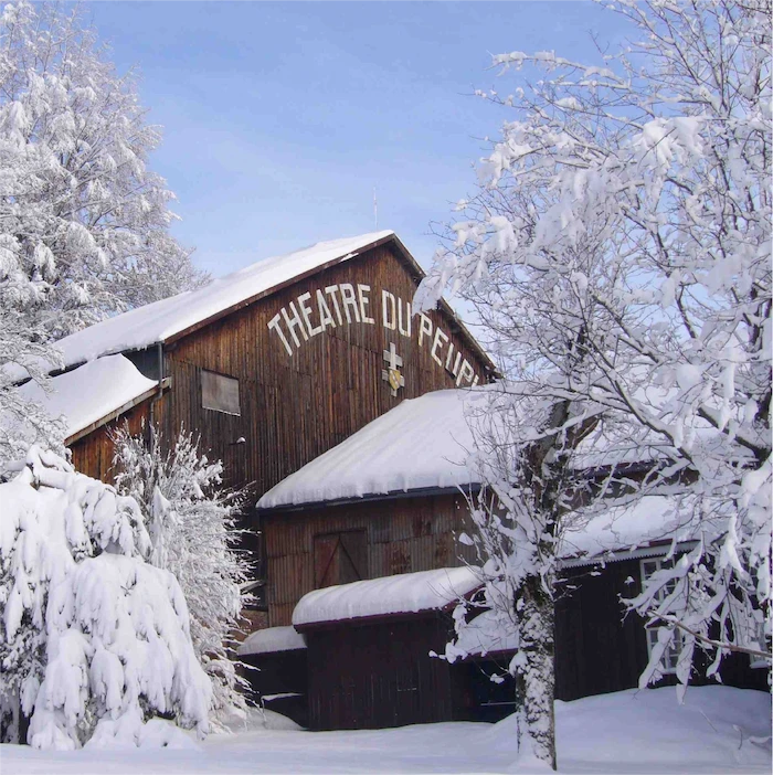 Wooden building with 'THEATRE DU PEUPLE' sign, surrounded by snow-covered trees and ground under a clear blue sky.