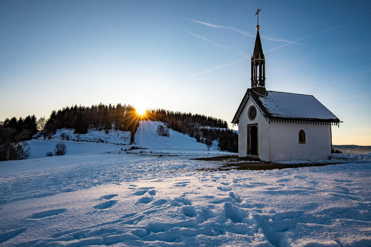 Small chapel with a steeple on a snow-covered field at sunset with sun rays peeking over a tree-covered hill.