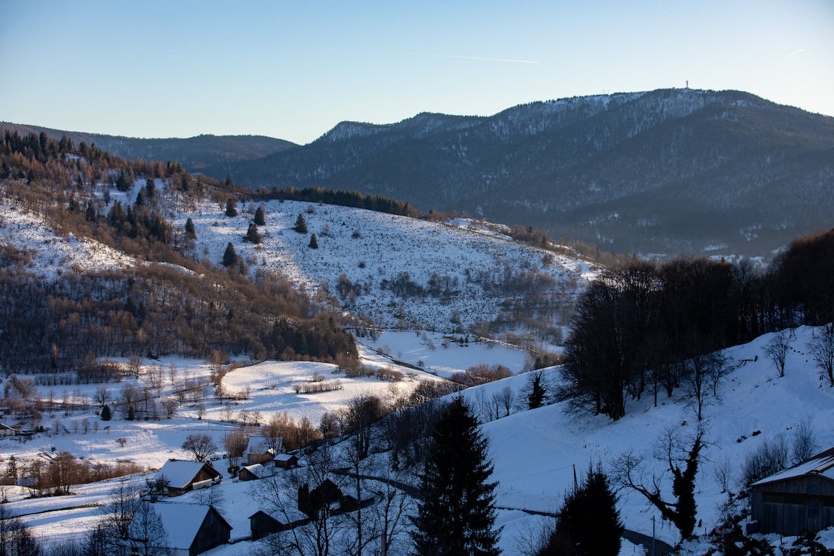 Snow-covered rural hillside with scattered trees and houses under a clear sky near forested mountains.