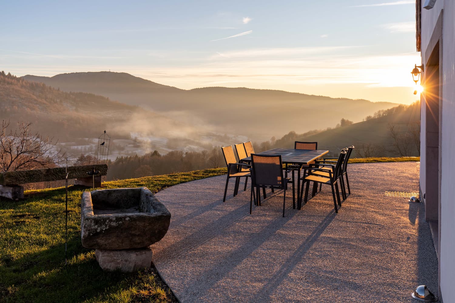 Snow-covered rural hillside with scattered trees and houses under a clear sky near forested mountains.
