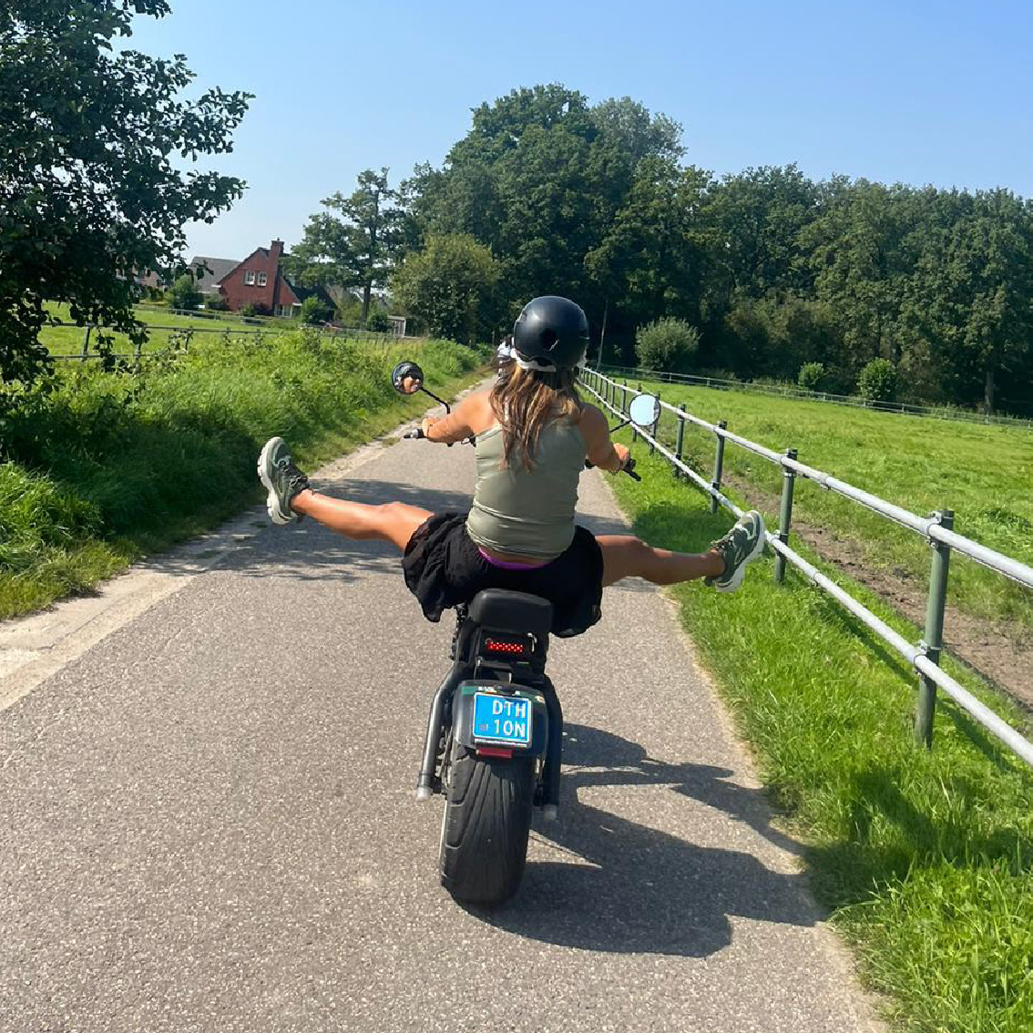 Persoon rijdt op een e-chopper over een landelijke weg, met het groene landschap en weilanden op de achtergrond, benen speels opzij gestrekt tijdens het rijden.