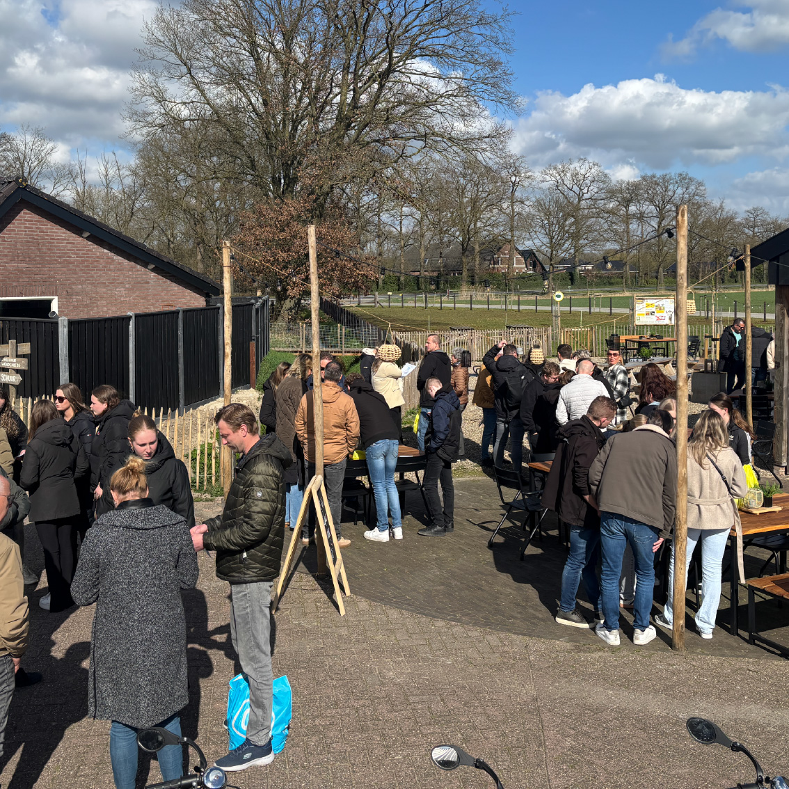 Groep mensen staat buiten op het erf van Er Was Eens Woudenberg bij picknicktafels, in gesprek en genietend van een gezamenlijke activiteit in een landelijke omgeving met boerderijen en bomen.
