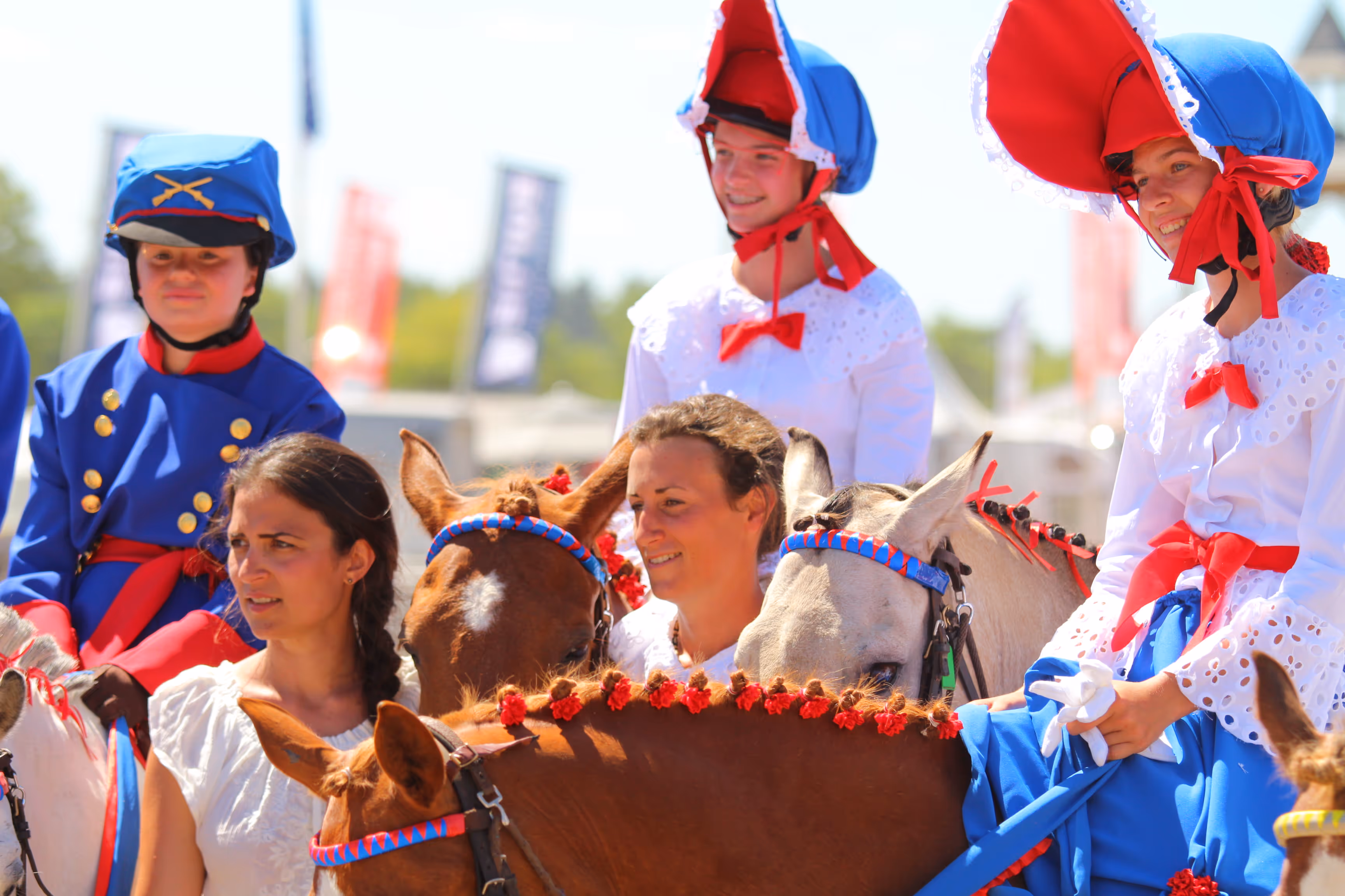 Group of people in traditional costumes, including blue outfits with red bows and bonnets, standing beside decorated horses.