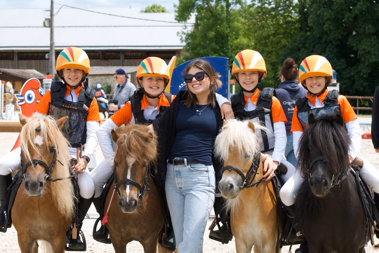 Four children in orange and white riding gear and helmets sitting on ponies, standing next to a woman in sunglasses and casual clothes smiling.