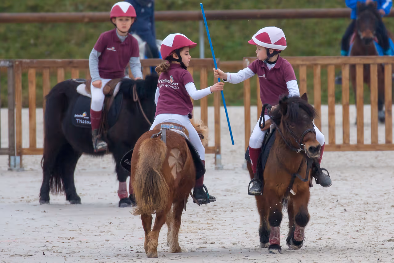 Two children in maroon helmets and shirts handing off a blue stick while riding ponies in an arena.