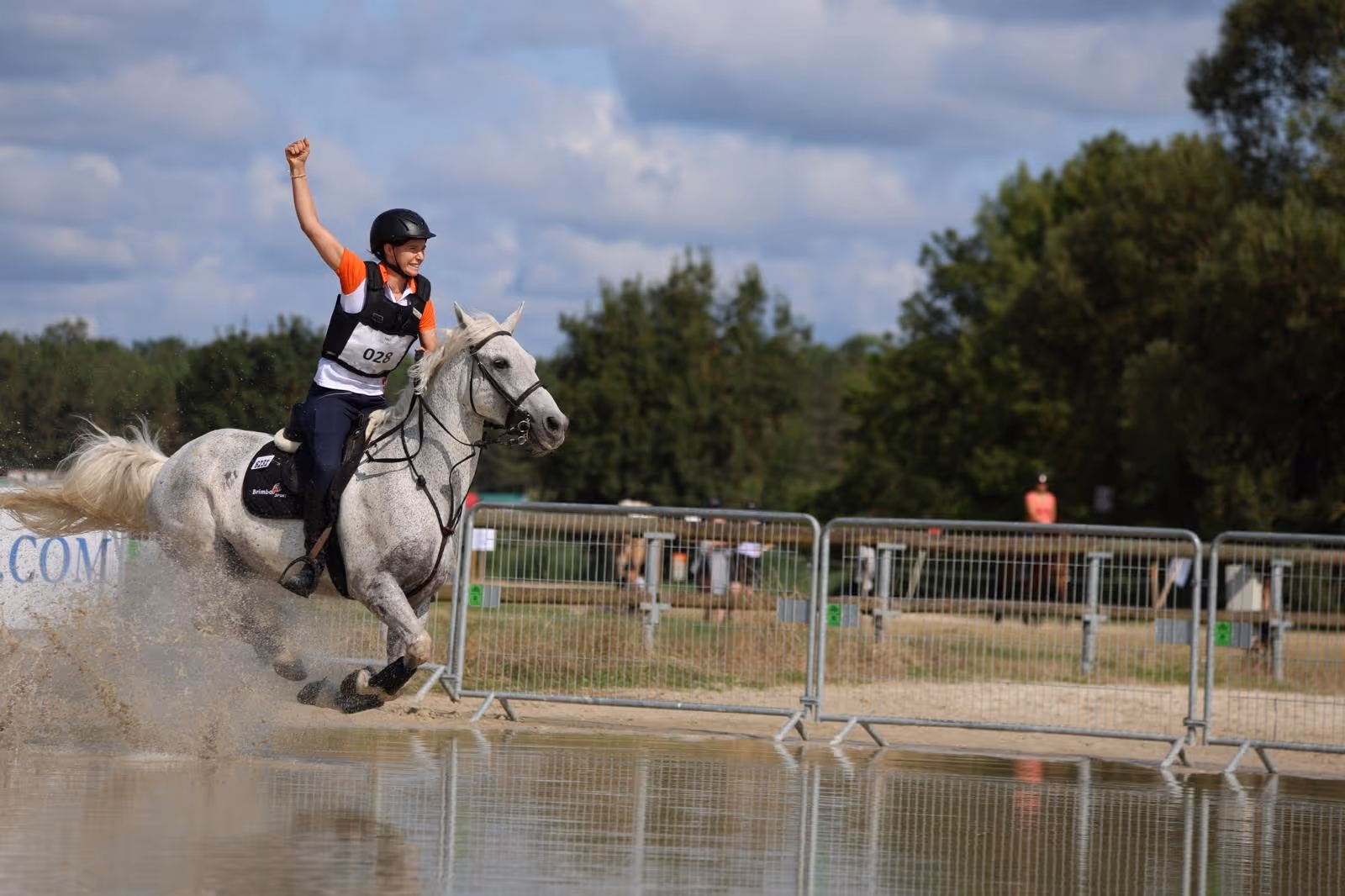 Equestrian rider in white pants and colorful helmet on a brown horse trotting across a field with green tents in the background.