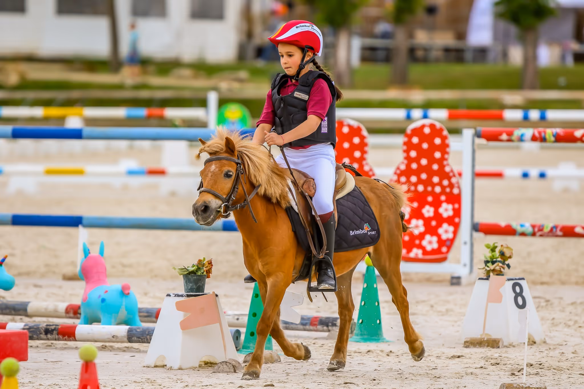 Young girl wearing red helmet and protective vest riding a pony in an equestrian arena with colorful obstacles.