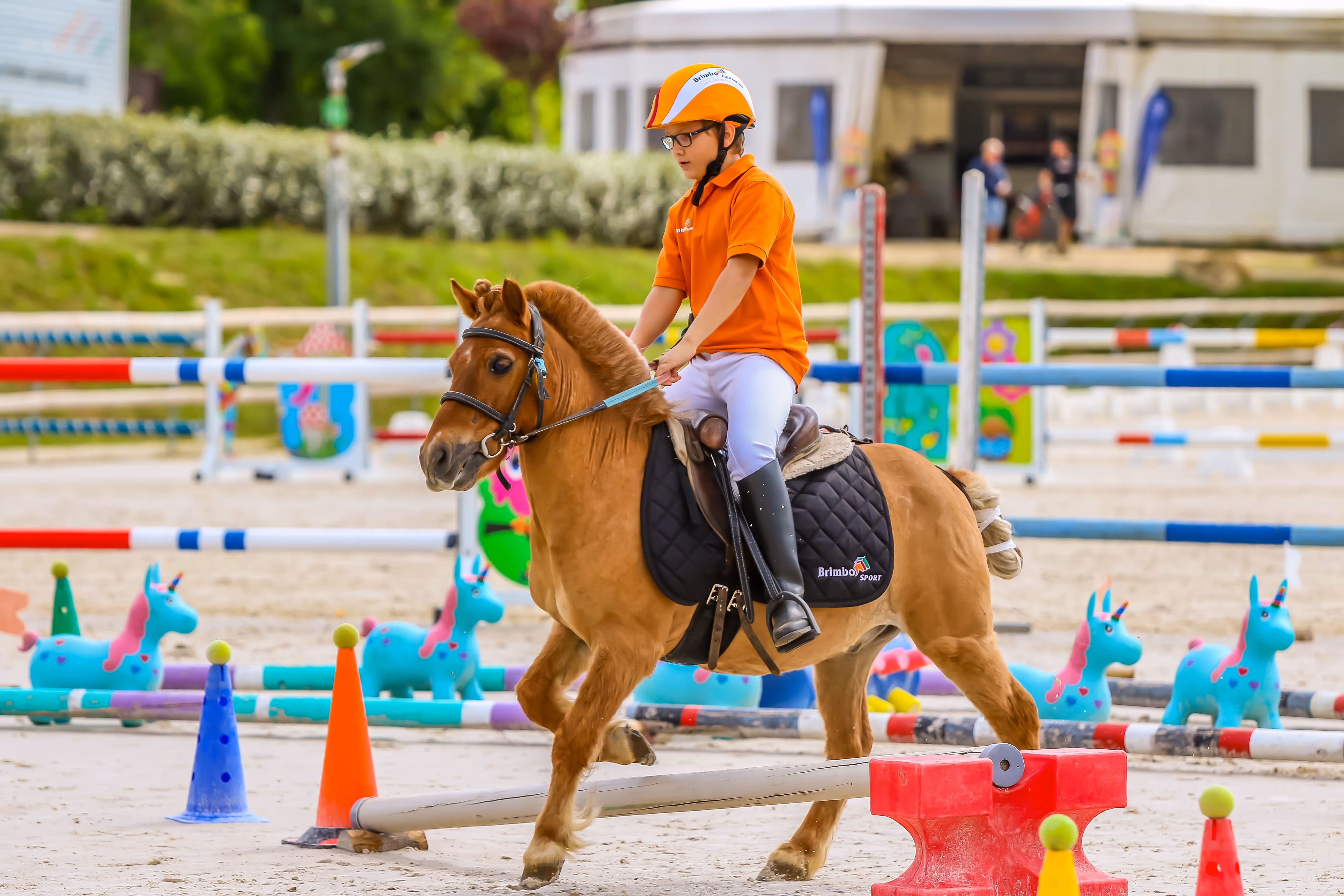 Young rider in orange shirt and helmet riding a pony over a low jump in an equestrian arena with colorful obstacles.