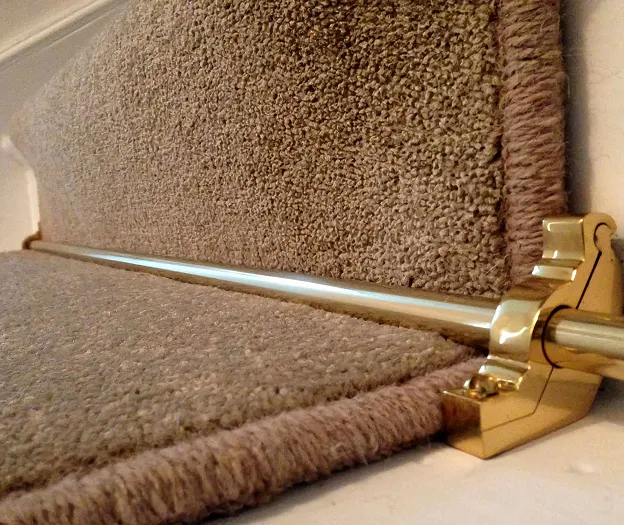 A close up of a brown carpet on stairs with bronze stair rods.