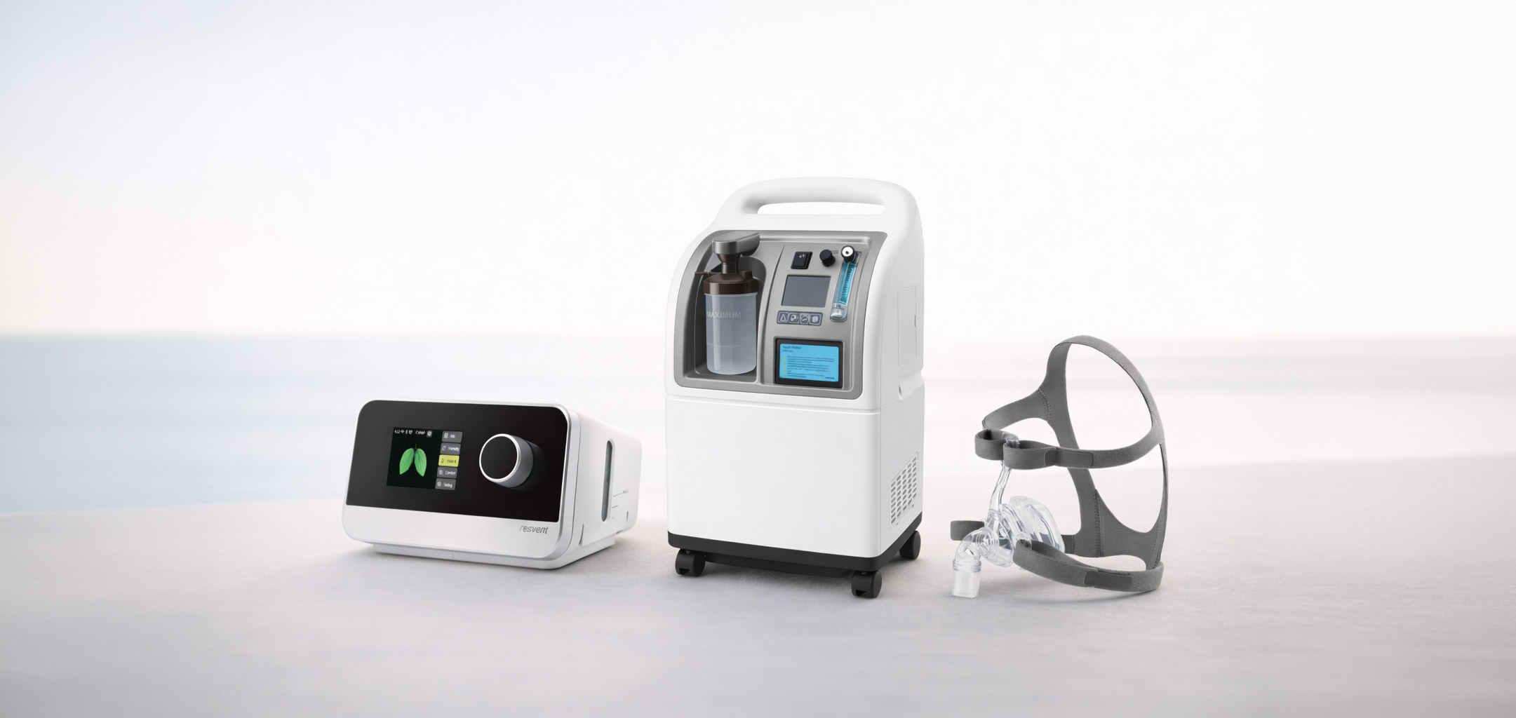Three medical respiratory devices on a plain surface: a white CPAP machine with a digital screen showing lungs, a white oxygen concentrator with control panel and humidifier bottle, and a gray headgear mask.
