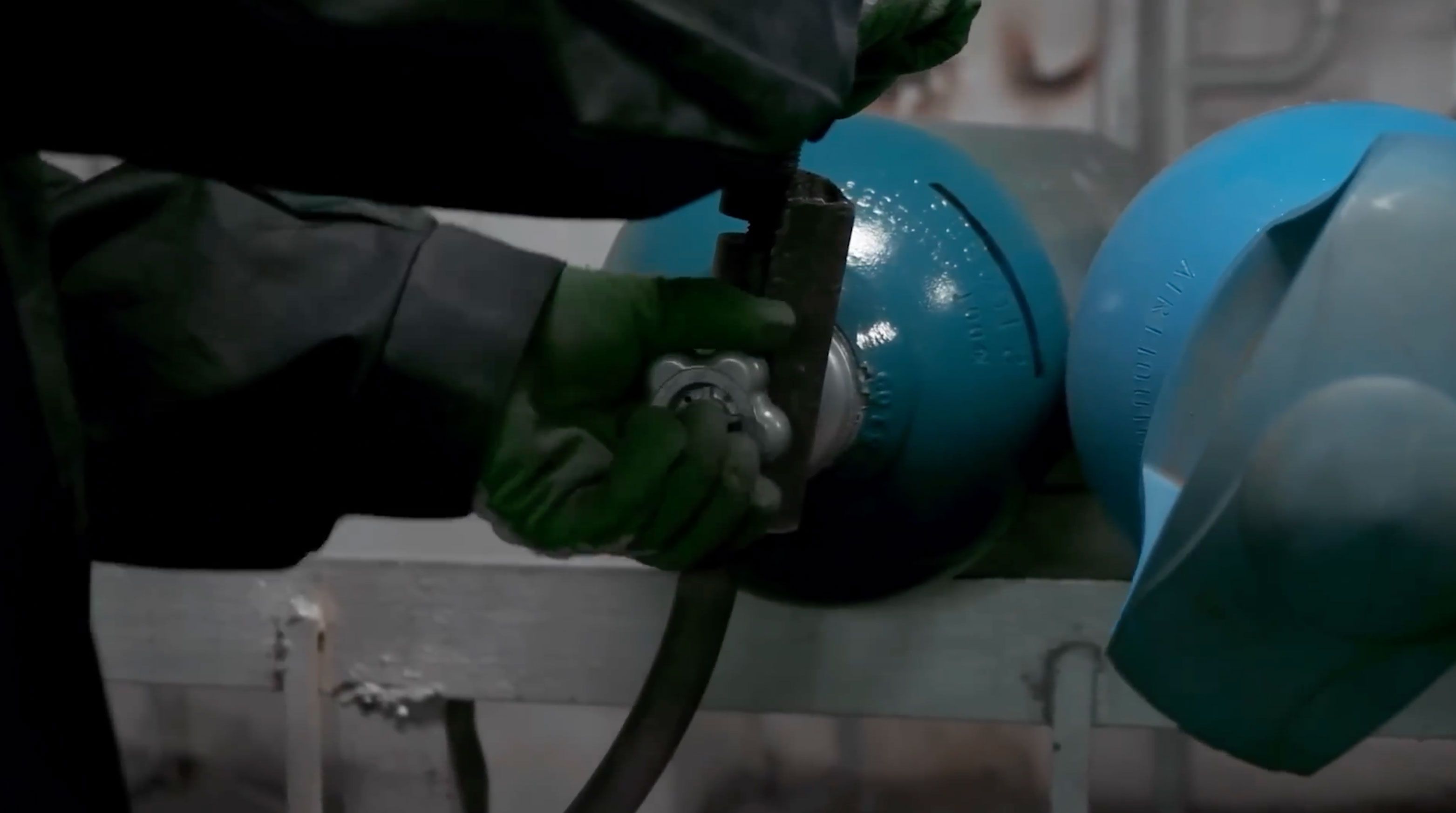 Person wearing green gloves adjusting a valve on a blue compressed gas cylinder.