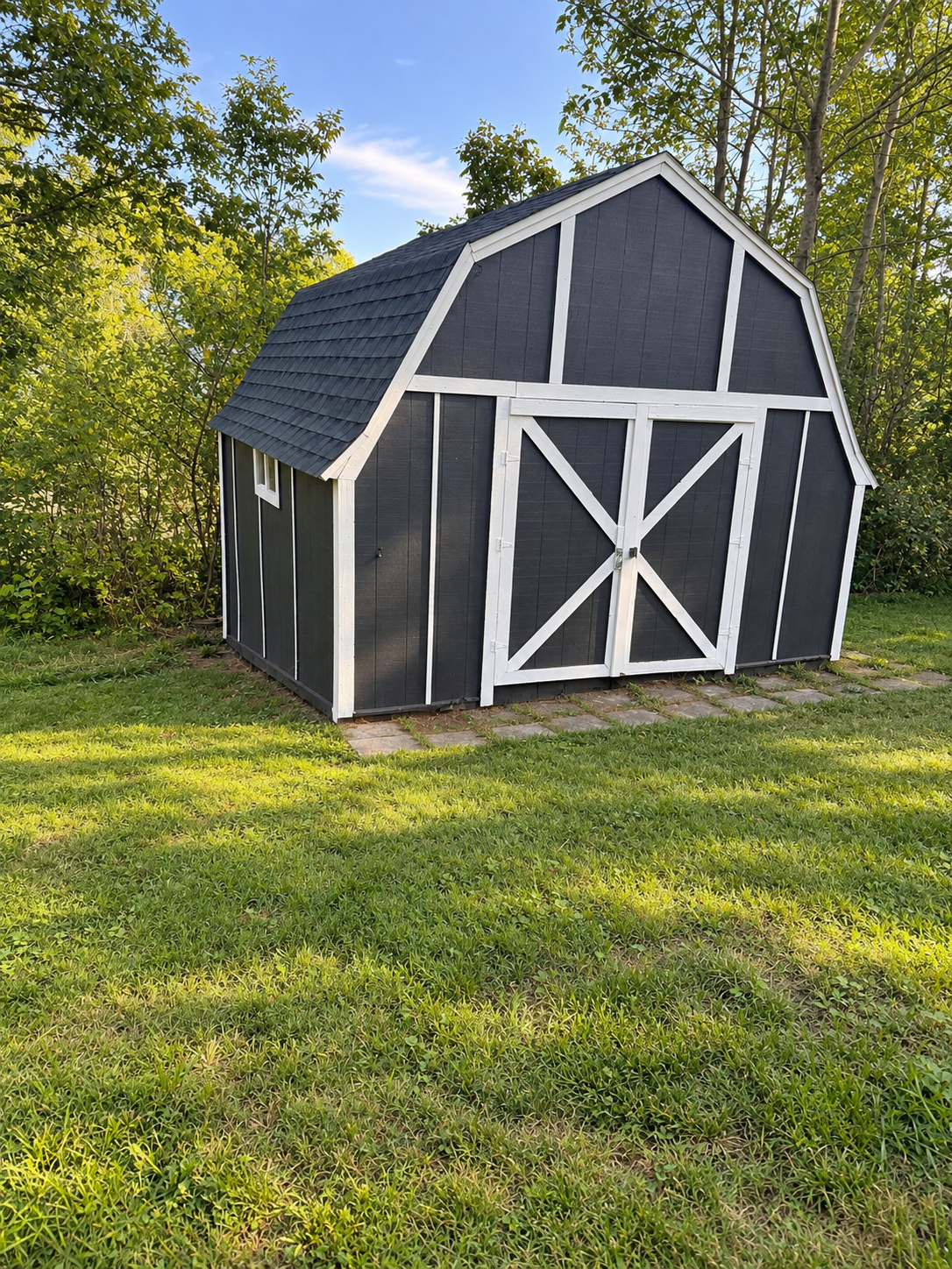 Barn-style storage shed painted charcoal gray with white trim, white cross-brace double doors, and a new dark asphalt shingle roof, set against trees