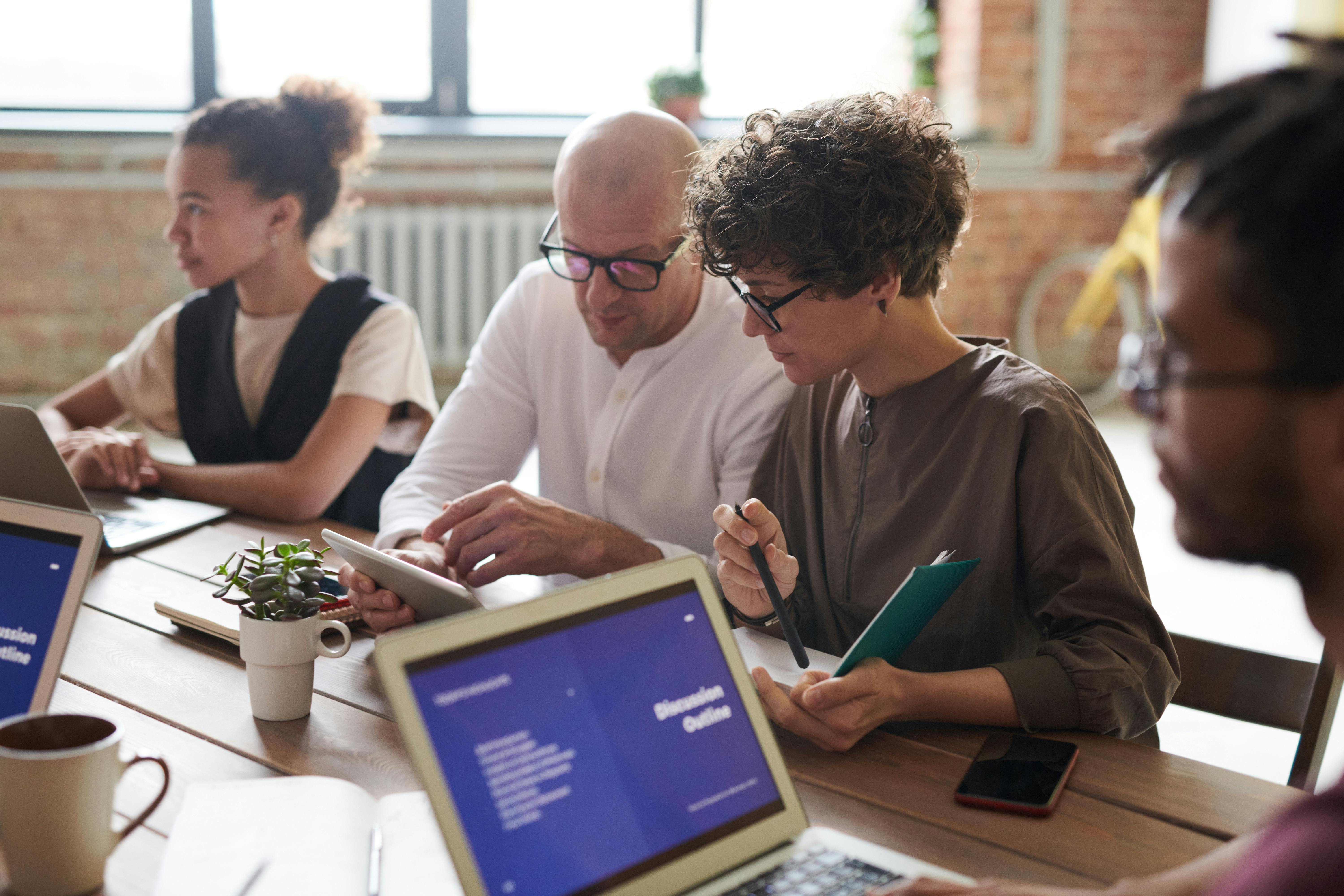 A group of colleagues sit around a table with their laptops and tablets. Two of the people are looking at a tablet together. 