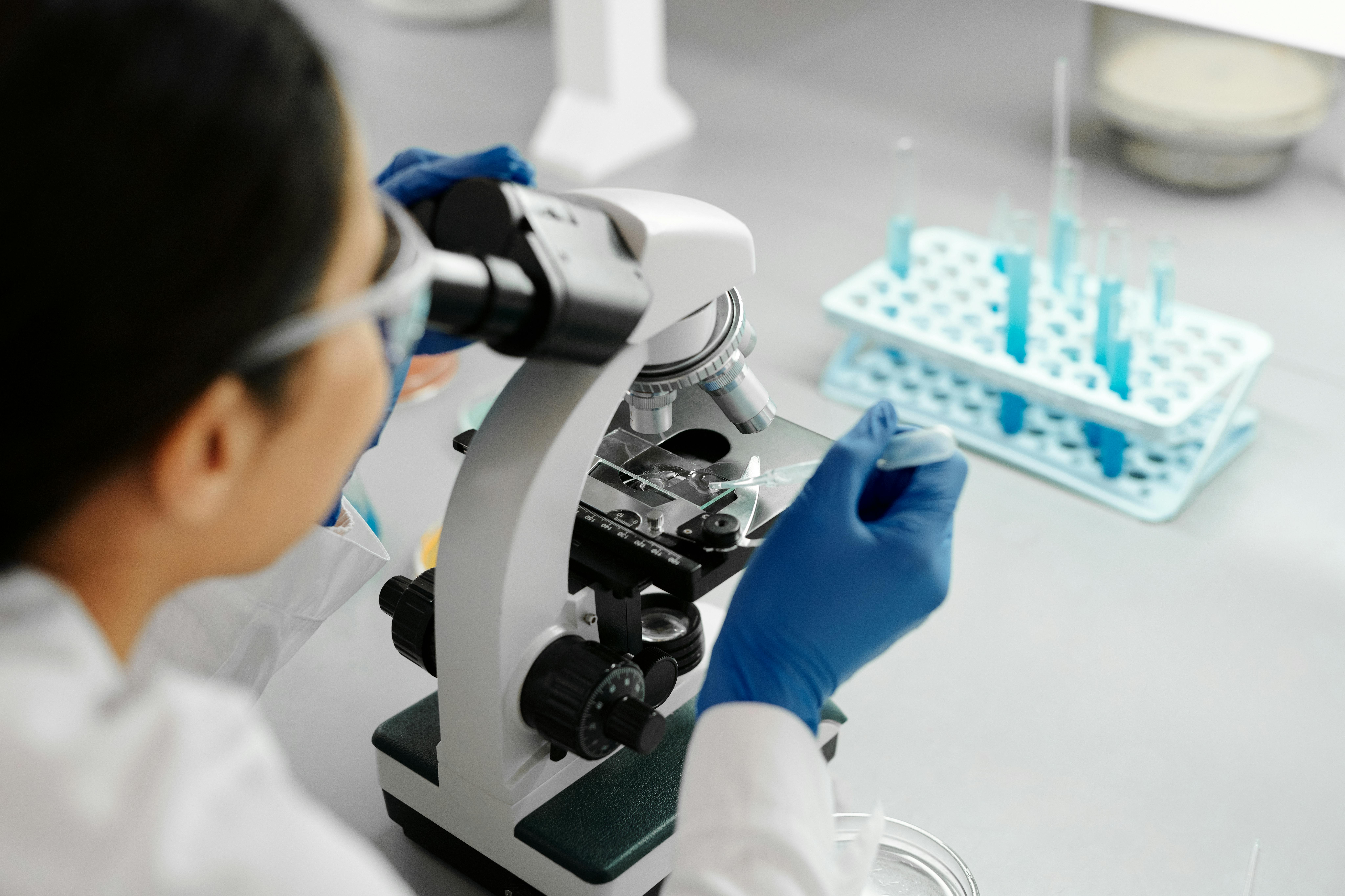 A researcher looks through a microscope in a sterile lab environment. There's a stand with test tubes on the table in front of them. 