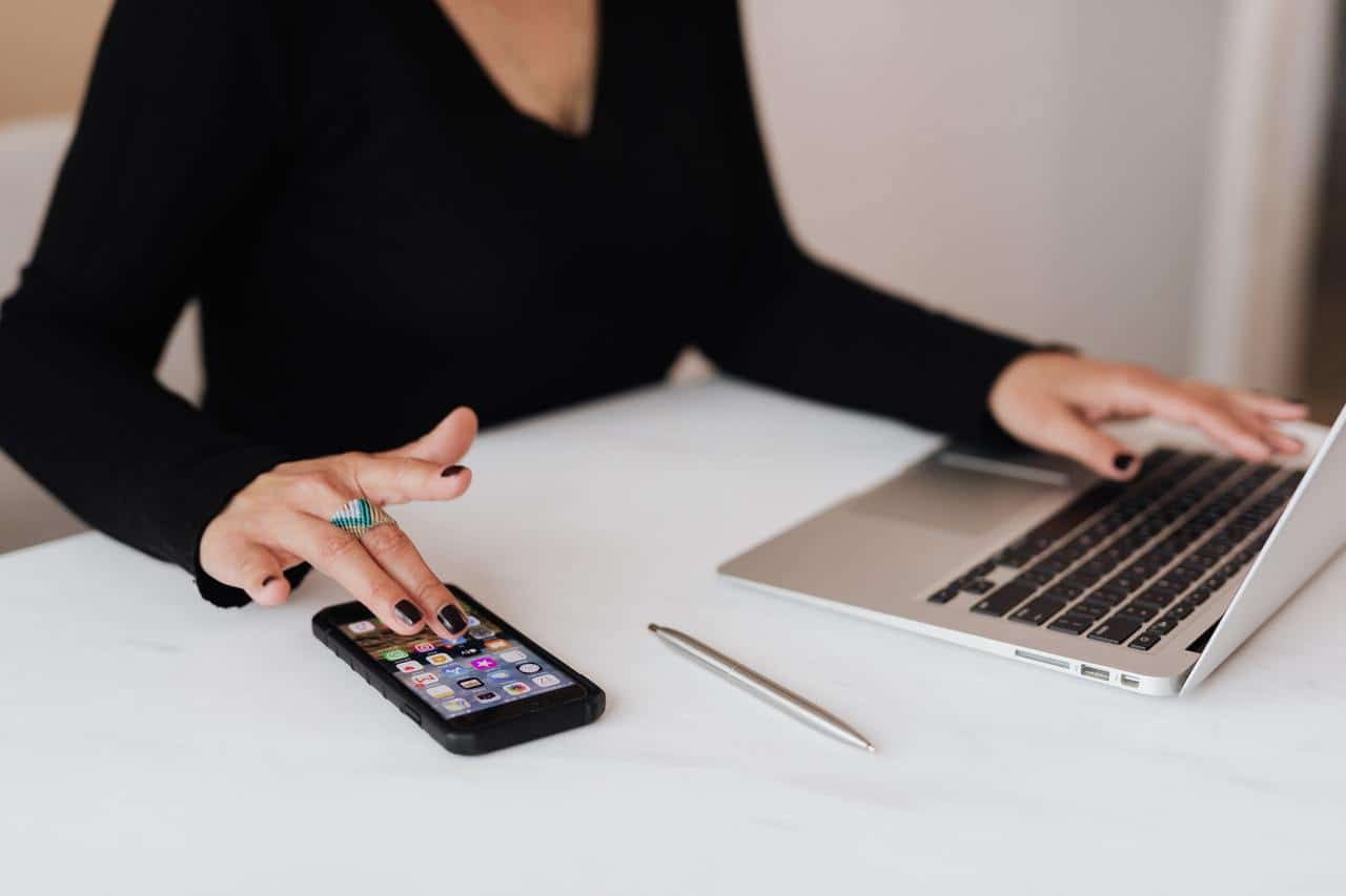 A person's hands typing on a laptop, with a phone showing the Instagram app nearby.