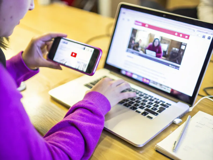 Hands holding a phone recording a YouTube video playing on a laptop.