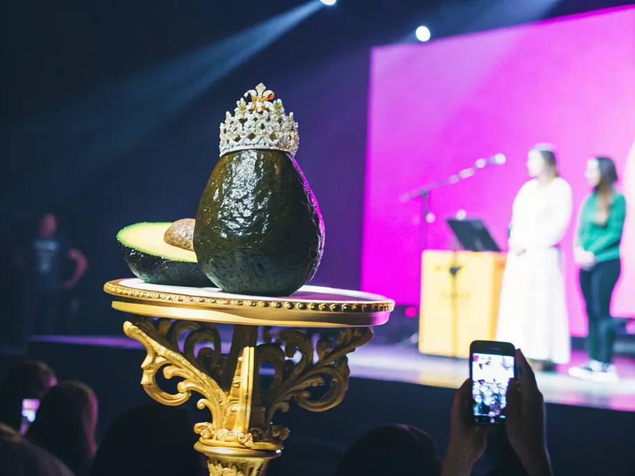 A single avocado crowned with sesame seeds and microgreens on a golden pedestal, surrounded by people taking photos.