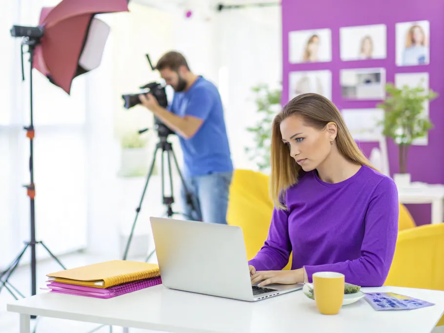 An influencer focused on creating content at a minimalist desk while a photographer sets up a shoot in the background.