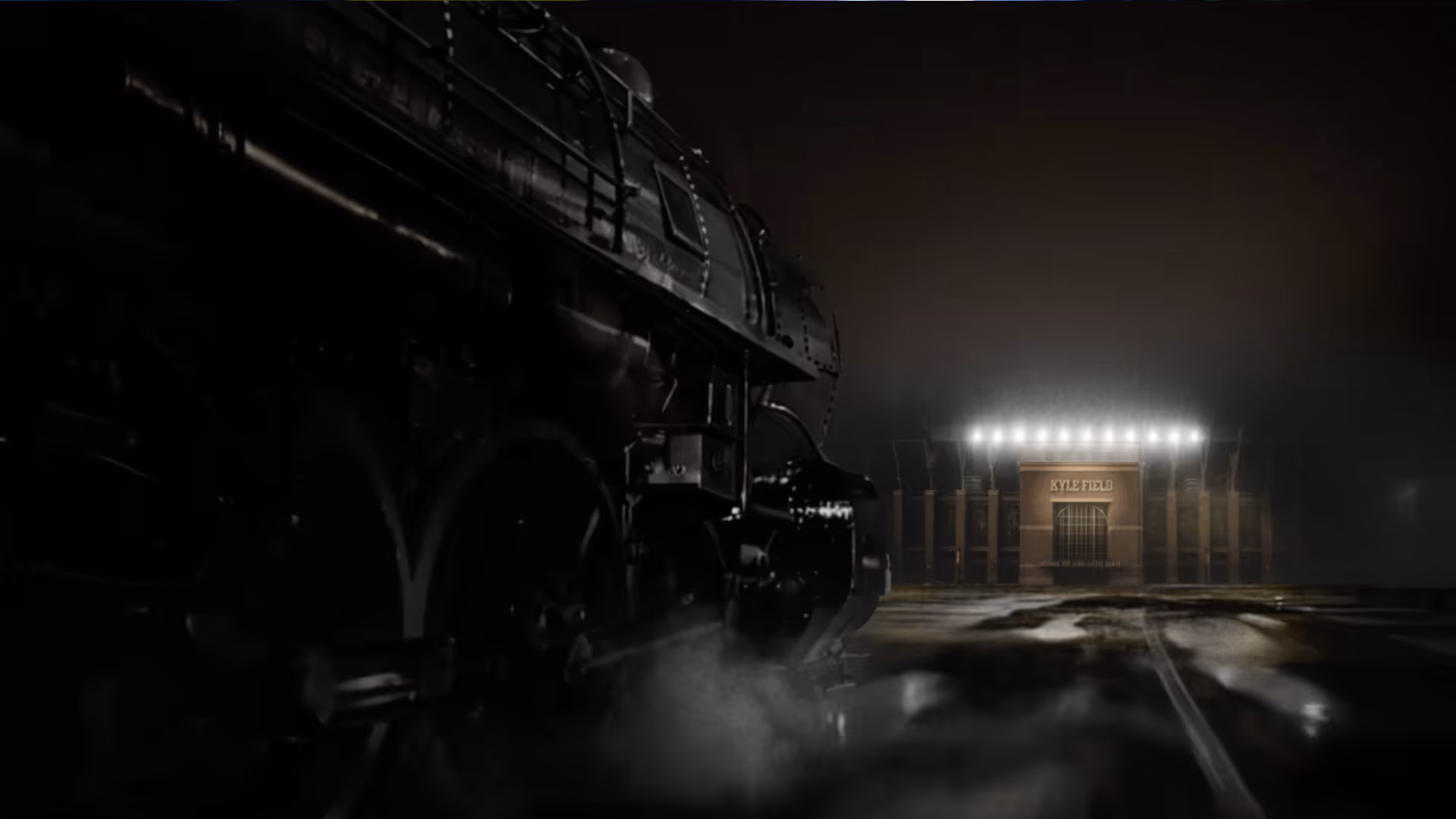 Side view of a black steam locomotive at night with steam rising, facing an illuminated stadium entrance labeled Kyle Field.
