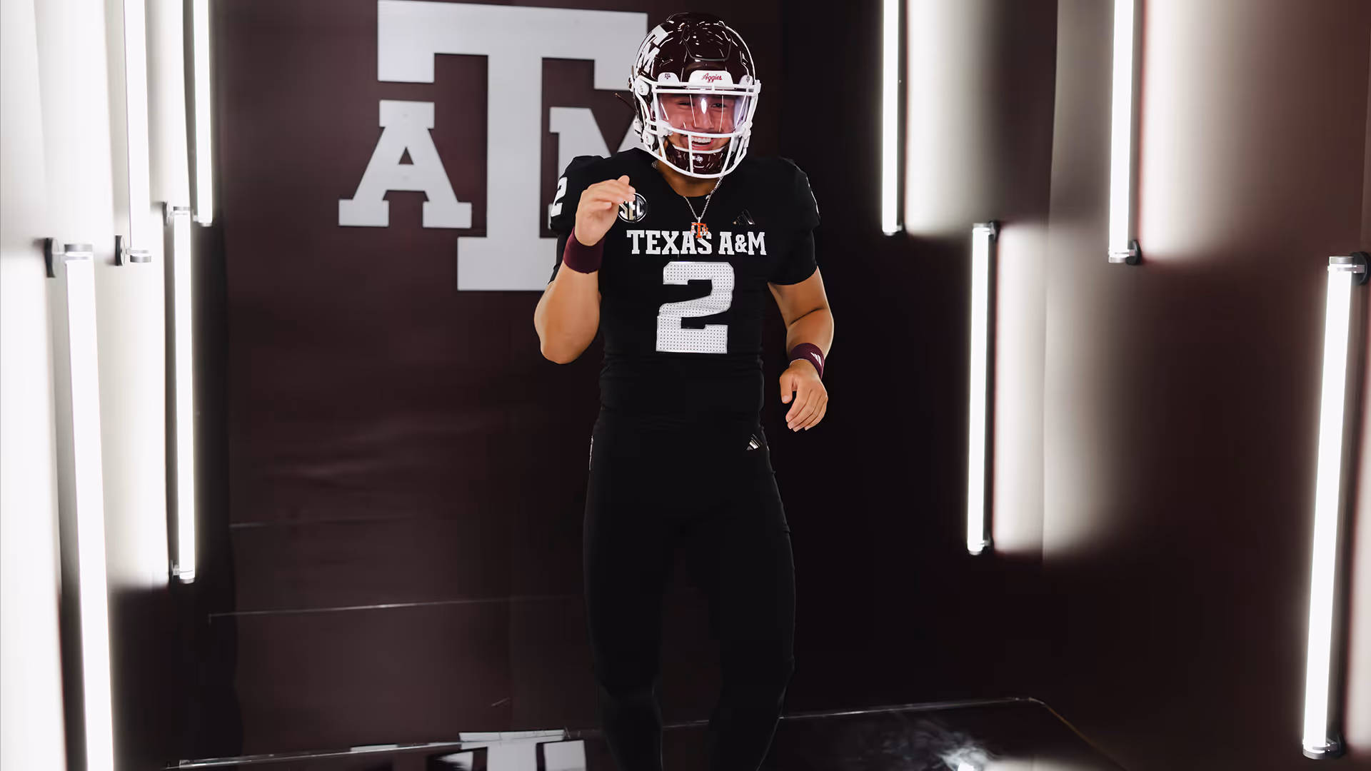 Football player wearing black Texas A&M jersey number 2 and helmet, standing in a dimly lit hallway with illuminated vertical lights.