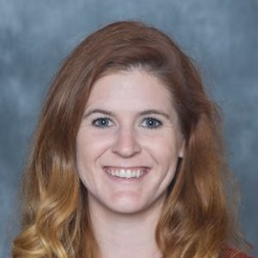 Smiling woman with long wavy red hair and blue eyes against a gray background.