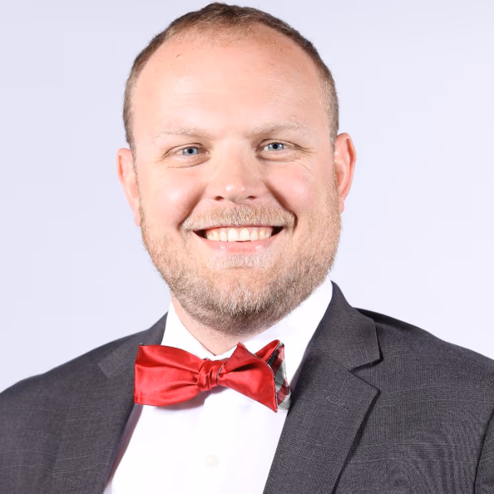 Smiling man wearing a dark suit jacket, white shirt, and red bow tie against a light background.
