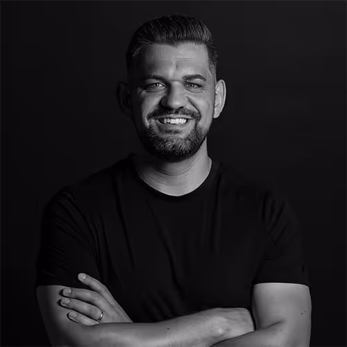 Smiling man with folded arms wearing a black t-shirt against a dark background.