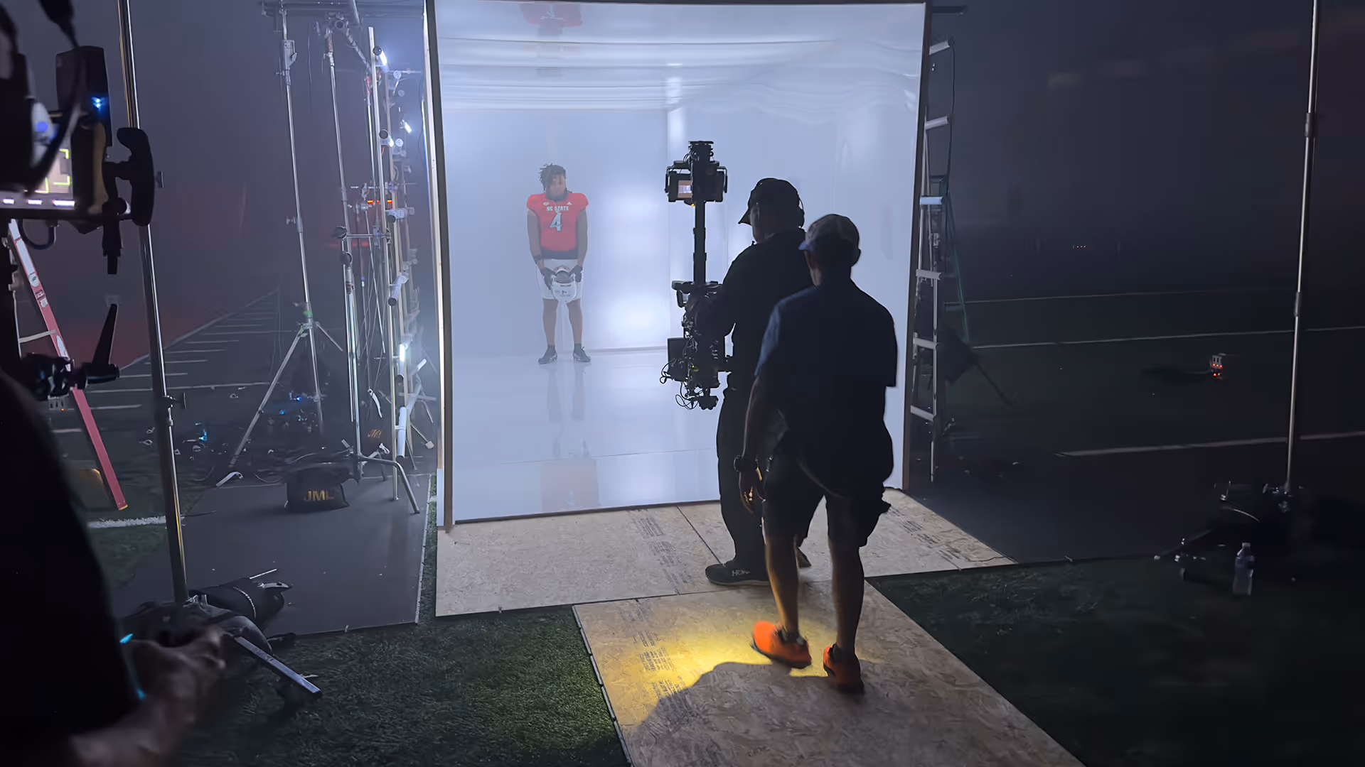 Film crew filming a football player in red jersey number 4 inside a lighted white set at night.