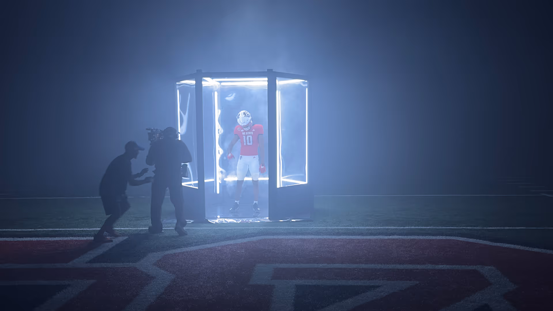 Football player wearing red NC State jersey number 10 stands inside a glowing transparent cube on a field while being filmed by a camera crew.