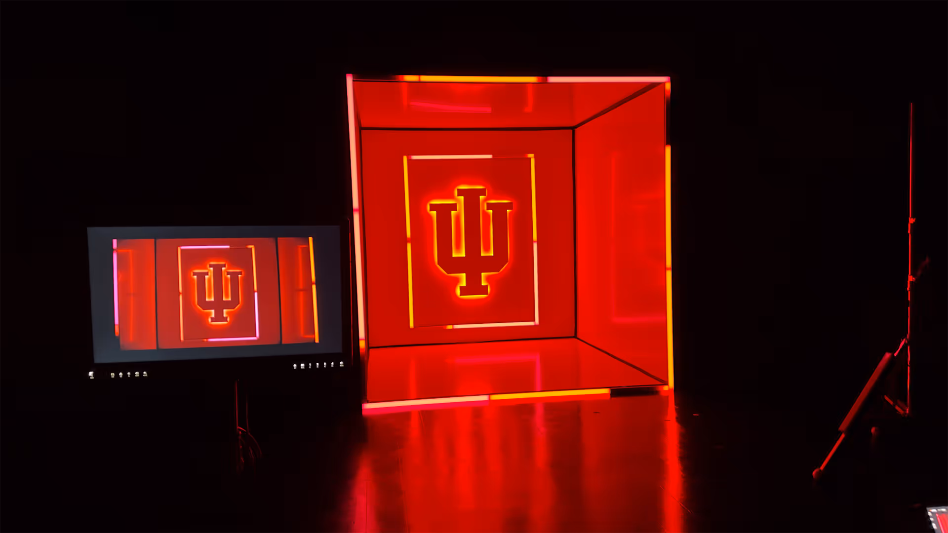 Illuminated red and orange Indiana University trident logo in a lighted box next to a monitor displaying the same image.