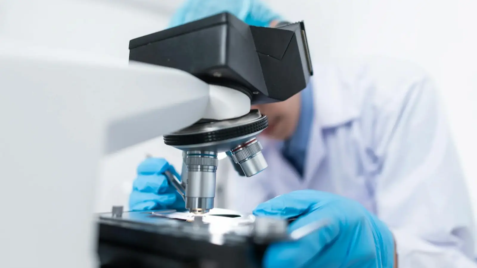 A worker in a lab coat and gloves looking into a microscope.