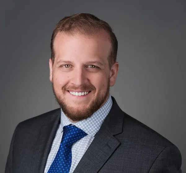 Smiling man with a beard wearing a dark suit, patterned white shirt, and blue tie against a gray background.