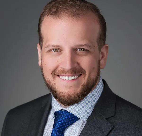 Smiling man with light brown hair, trimmed beard, wearing a dark suit, patterned shirt, and blue tie against a gray background.